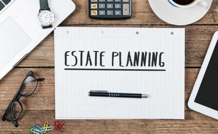Overhead shot features an estate planning document, set on a rustic wood desk among office essentials. A clean white laptop, calculator, glasses, pen, watch, tablet, colorful paperclips, and a steaming mug of coffee surround the document.