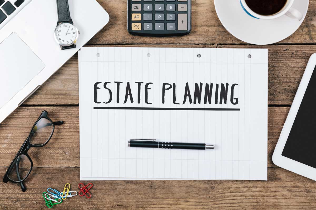 Overhead shot features an estate planning document, set on a rustic wood desk among office essentials. A clean white laptop, calculator, glasses, pen, watch, tablet, colorful paperclips, and a steaming mug of coffee surround the document.
